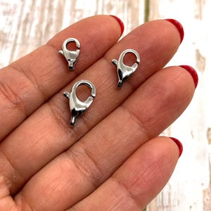 May include: Three shiny silver-colored lobster claw clasps. These clasps are commonly used in jewelry making and crafting. The clasps are shown against a background of a hand with red nail polish.