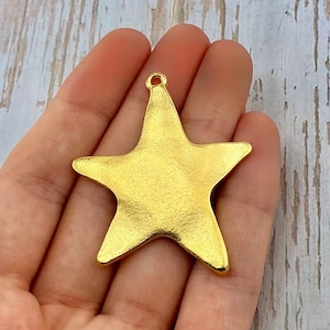 May include: A gold-colored star-shaped charm with a textured surface. The charm has five points and a small loop at the top for attaching to a necklace or bracelet. This charm is suitable for jewelry making.