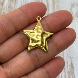 May include: A gold-colored star-shaped charm with a textured surface. The charm has a small loop at the top for attaching to a necklace or bracelet. The star is approximately 1 inch wide.