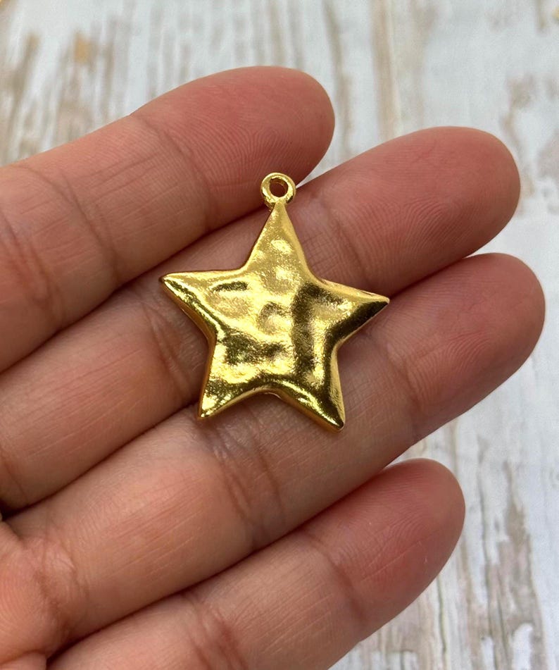 May include: A gold-colored star-shaped charm with a textured surface. The charm has a small loop at the top for attaching to a necklace or bracelet. The star is approximately 1 inch wide.