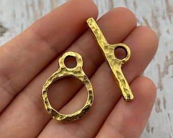May include: Two gold-colored toggle clasps. One is a ring with a hammered texture, and the other is a bar with a loop. The clasps are shown against a light-colored background.