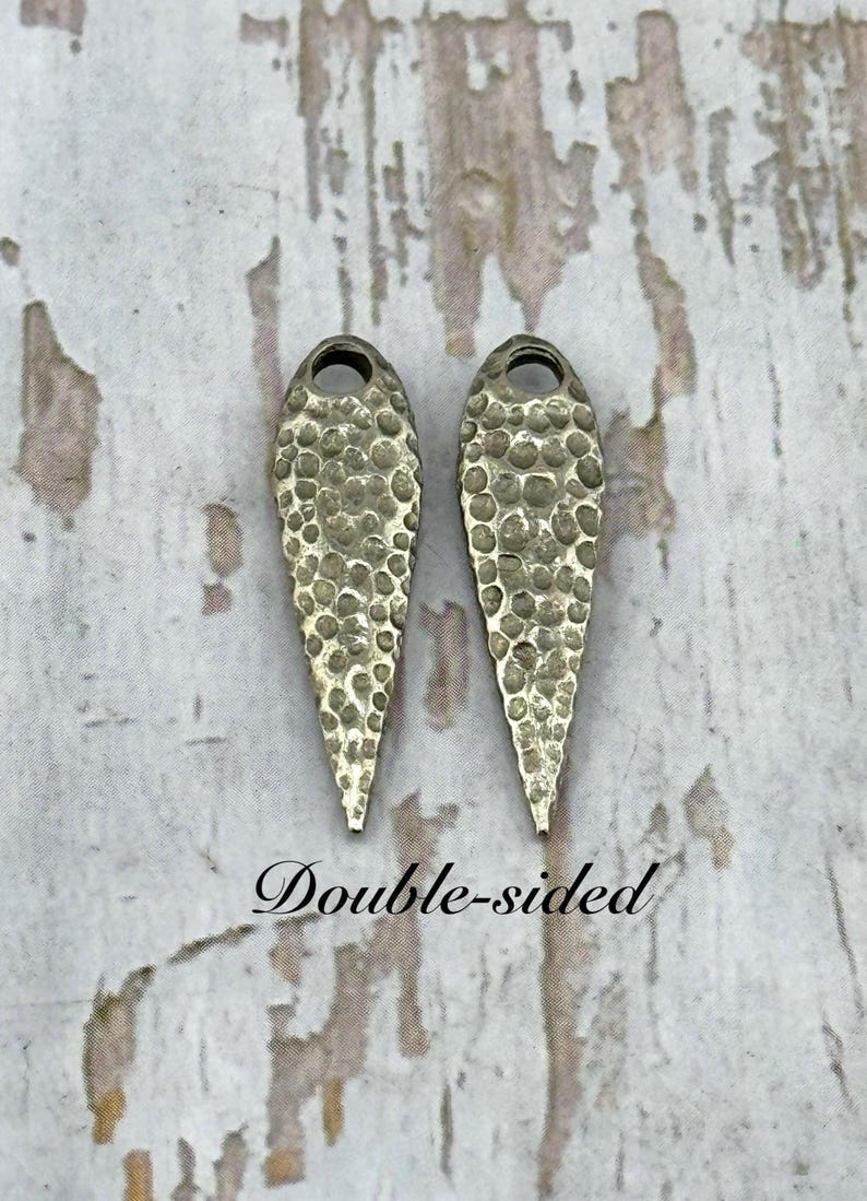 Pu&ograve; includere: Due orecchini bifacciali color argento con una texture martellata. Ogni orecchino ha la forma di una goccia con un piccolo foro nella parte superiore. Il testo "Double-sided" &egrave; scritto in corsivo sotto.