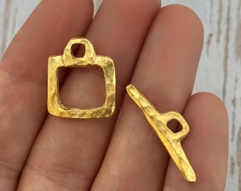 May include: Two gold-colored, hammered-texture jewelry clasps. One is a square shape with a loop at the top, and the other is a bar shape with a loop at one end. The clasps are likely for necklaces or bracelets.