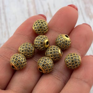 May include: Close-up of several gold-colored, round beads with a grid-like pattern of small, dark stones. The beads have a metallic sheen and a hole running through the center. The beads are likely for jewelry making.