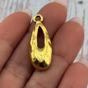 May include: A gold-colored pendant with a teardrop shape and a central cutout. The pendant has a textured surface and a small loop at the top for attaching to a chain or cord. Jewelry.