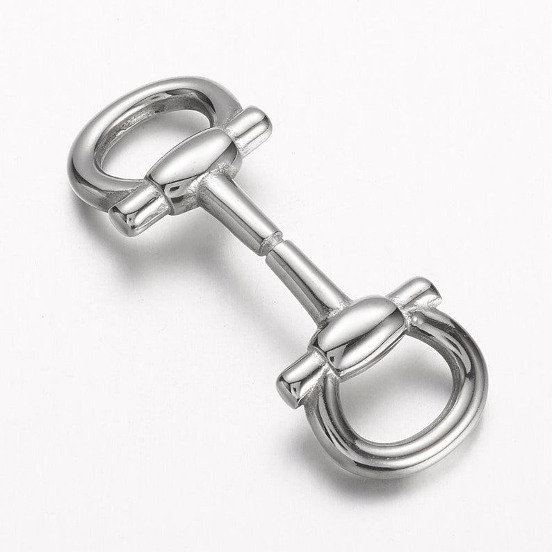 May include: A shiny, silver-colored horse bit with a central bar and rounded rings on each end. The bit has a polished, reflective surface, and is designed for use in equestrian activities. The design is symmetrical and detailed, with smooth curves and a classic shape.