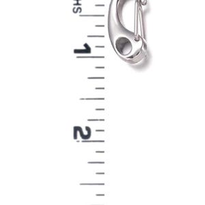May include: A silver-colored carabiner clip with a spring-loaded gate. The clip is approximately 3 inches long. The image includes a ruler for scale, showing the clip's size.