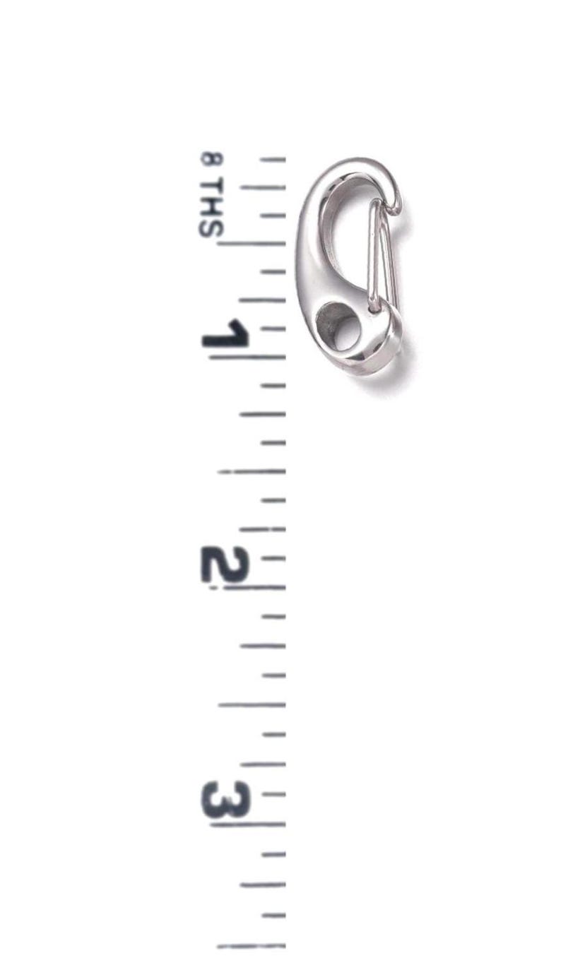 May include: A silver-colored carabiner clip with a spring-loaded gate. The clip is approximately 3 inches long. The image includes a ruler for scale, showing the clip's size.