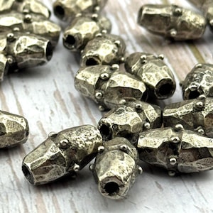 May include: Close-up of several silver-colored, barrel-shaped beads. Each bead has a hammered texture and small, round accents. The beads are scattered, with some in focus and others blurred, suggesting a craft or jewelry-making theme.