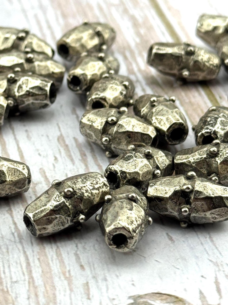 May include: Close-up of several silver-colored, barrel-shaped beads. Each bead has a hammered texture and small, round accents. The beads are scattered, with some in focus and others blurred, suggesting a craft or jewelry-making theme.