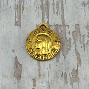 May include: A gold-colored pendant with the word "MARIE" and a profile of a woman's face. The pendant has a loop at the top for a chain and is surrounded by a circular border with cross symbols.