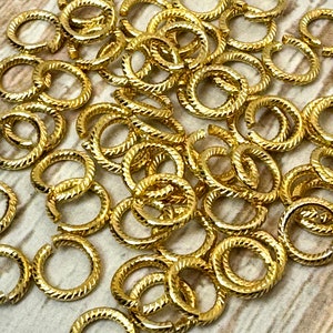 May include: A collection of small, gold-colored jump rings with a textured, twisted rope design. These jewelry findings are open and ready to be used in various crafting projects. The rings are scattered across a light-colored surface.