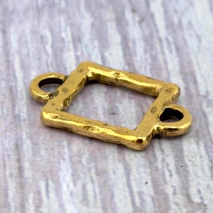 May include: A gold-toned metal jewelry connector featuring a rectangular frame with a textured, hammered surface. The piece includes two small circular loops extending from opposite sides of the frame, designed for attaching chains or other components.
