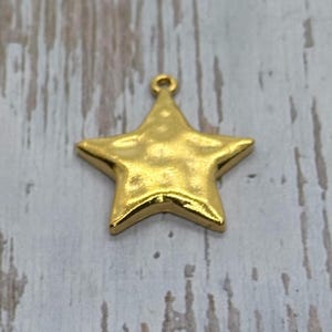 May include: A gold-colored star-shaped charm with a textured surface and a small loop at the top for hanging. The charm is a bright, shiny gold color and is likely made of metal.