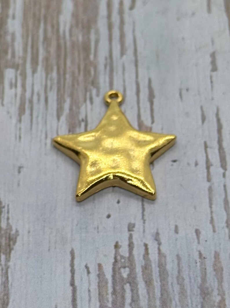 May include: A gold-colored star-shaped charm with a textured surface and a small loop at the top for hanging. The charm is a bright, shiny gold color and is likely made of metal.