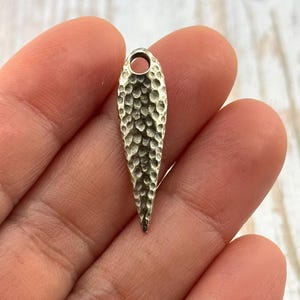 Pu&ograve; includere: Un ciondolo a forma di goccia, color argento, con texture martellata e un piccolo foro nella parte superiore. Il ciondolo &egrave; tenuto in mano da una persona. La superficie del ciondolo ha una texture a fossette.