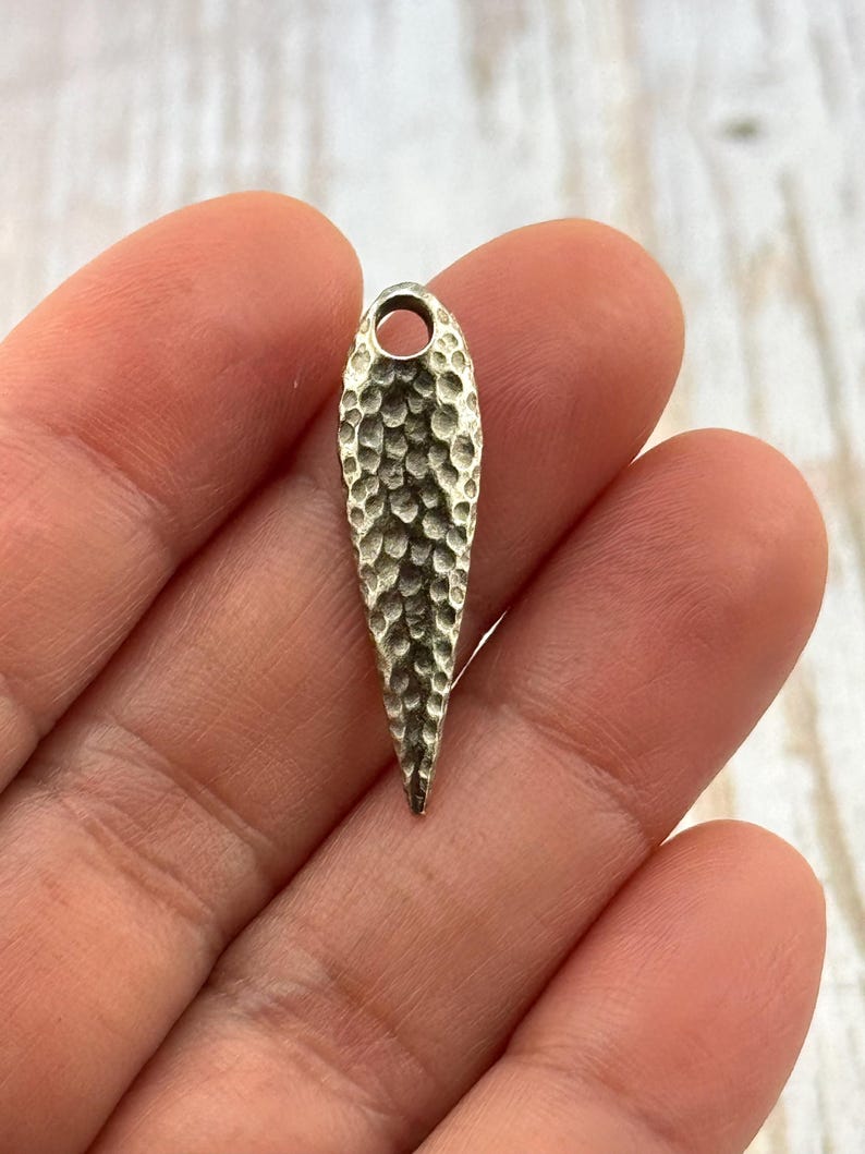 Pu&ograve; includere: Un ciondolo a forma di goccia, color argento, con texture martellata e un piccolo foro nella parte superiore. Il ciondolo &egrave; tenuto in mano da una persona. La superficie del ciondolo ha una texture a fossette.