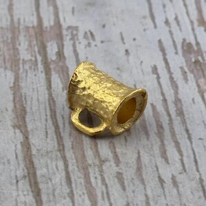 Puede incluir: Un peque&ntilde;o dije de taza de color dorado, con una superficie texturizada y un asa. El dije probablemente est&aacute; hecho de metal y est&aacute; dise&ntilde;ado para parecerse a una jarra de cerveza en miniatura. El dije se muestra sobre una superficie de madera de color claro.