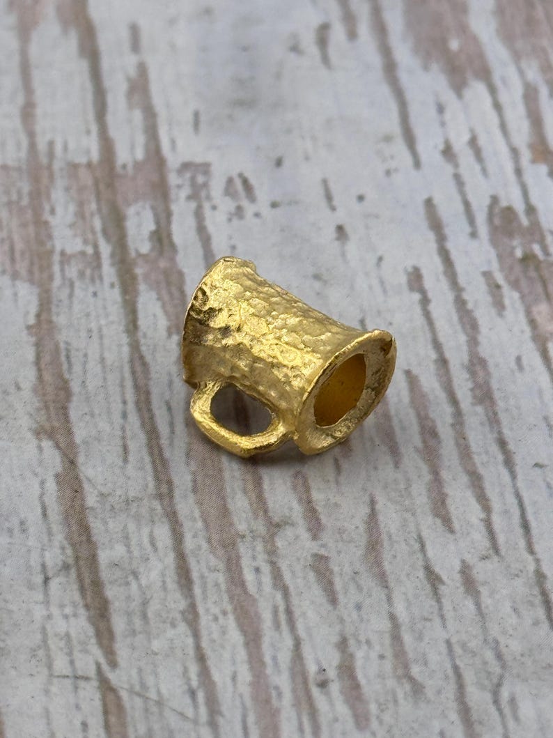 Puede incluir: Un peque&ntilde;o dije de taza de color dorado, con una superficie texturizada y un asa. El dije probablemente est&aacute; hecho de metal y est&aacute; dise&ntilde;ado para parecerse a una jarra de cerveza en miniatura. El dije se muestra sobre una superficie de madera de color claro.