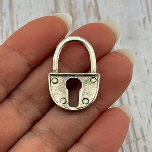 May include: A small, silver-colored padlock with a curved handle. The lock has a keyhole and is secured with rivets. The padlock is held in a hand, suggesting its small size and potential use in jewelry or crafts.