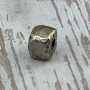 May include: A small, silver-colored, cube-shaped bead with a slightly irregular surface. The bead has a hole drilled through it, suggesting it is for stringing or jewelry making. The bead's metallic appearance gives it a vintage or antique feel.