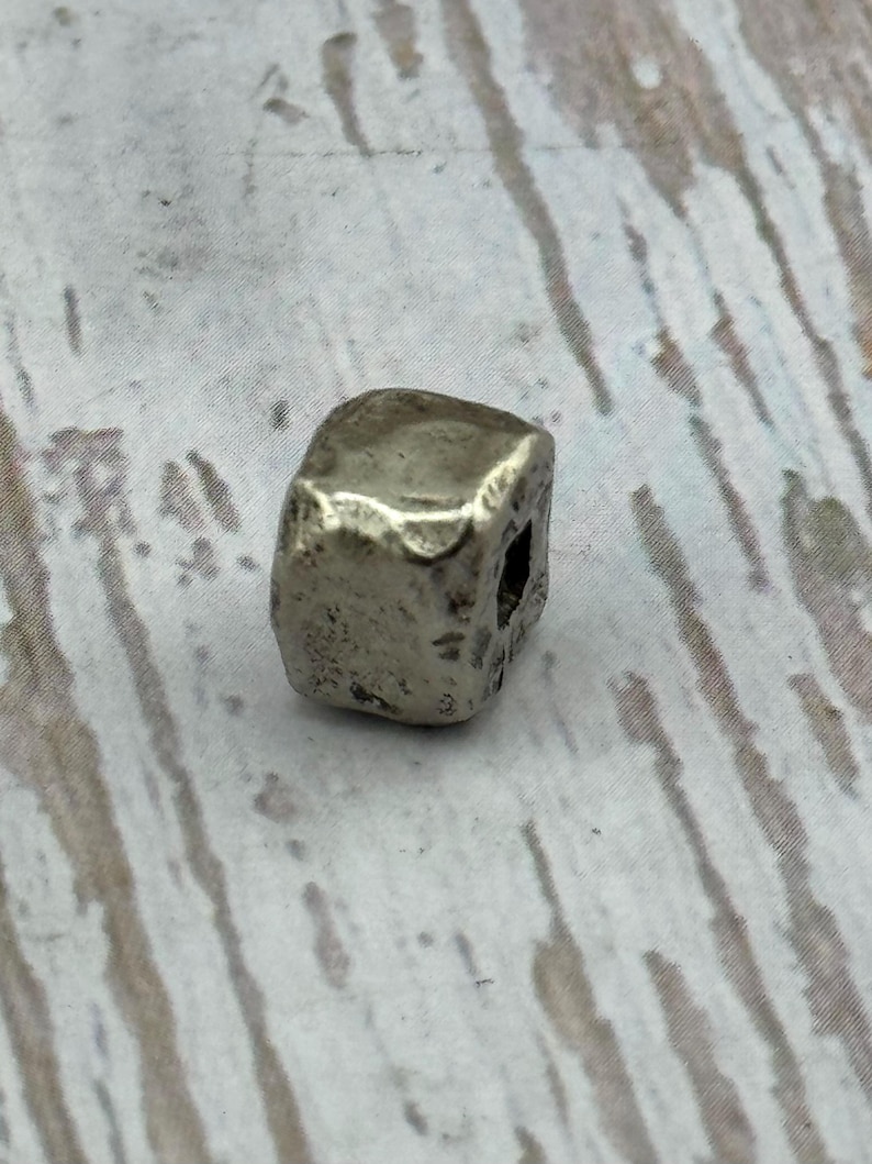 May include: A small, silver-colored, cube-shaped bead with a slightly irregular surface. The bead has a hole drilled through it, suggesting it is for stringing or jewelry making. The bead's metallic appearance gives it a vintage or antique feel.