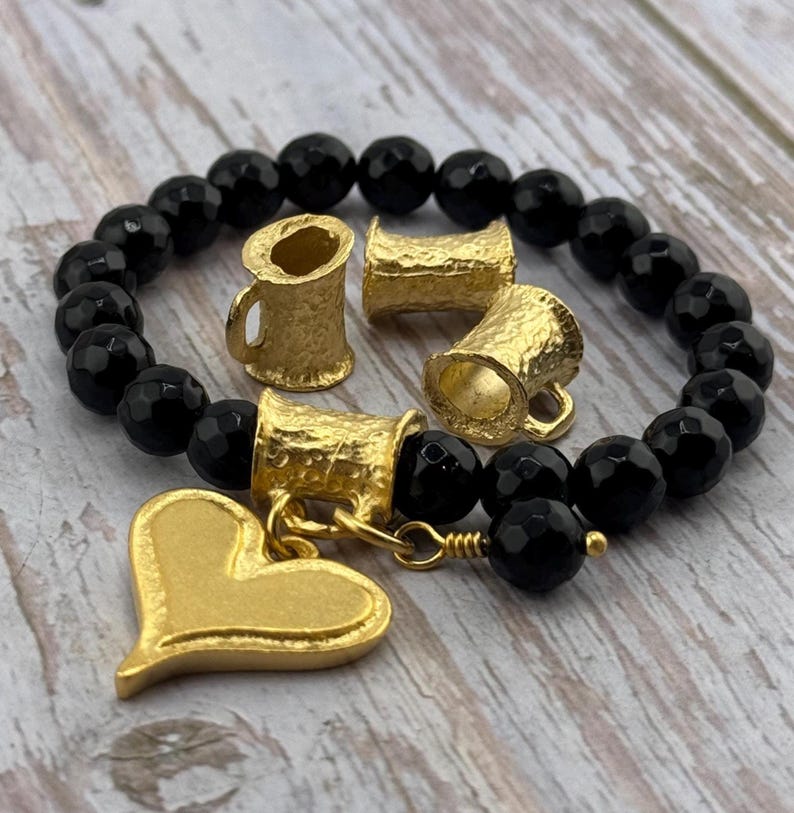 Puede incluir: Una pulsera de cuentas negras con detalles dorados. La pulsera presenta cuentas negras facetadas y elementos decorativos dorados, incluyendo peque&ntilde;os dijes en forma de taza y un dije en forma de coraz&oacute;n.
