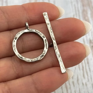 May include: A silver-toned toggle clasp set, featuring a textured, hammered finish. The set includes a circular ring with a loop and a straight bar with a small ball at one end. The clasp is designed for jewelry making and crafting.