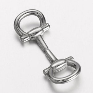 May include: A silver-colored metal horse bit with two rounded rings and a central bar. The bit has a polished finish and is designed for equestrian use. The image is taken from a high angle, showing the details of the bit.