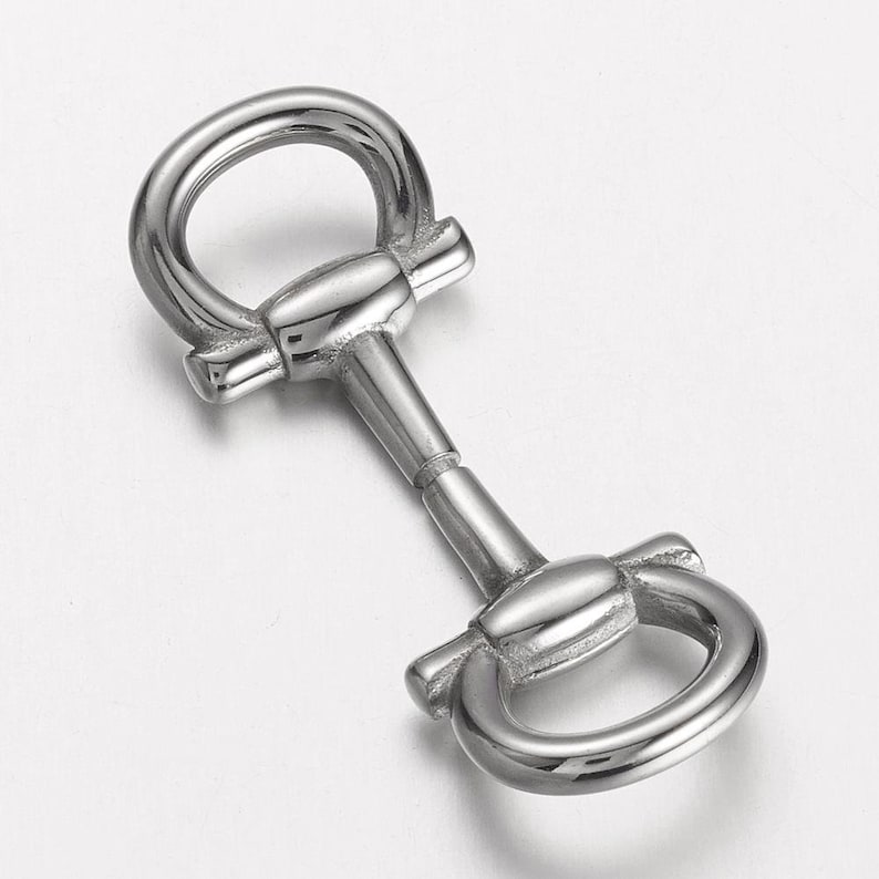 May include: A silver-colored metal horse bit with two rounded rings and a central bar. The bit has a polished finish and is designed for equestrian use. The image is taken from a high angle, showing the details of the bit.