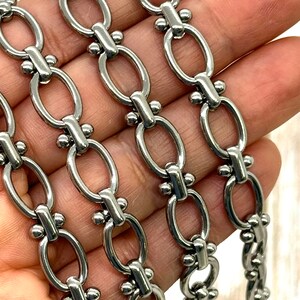 May include: Close-up of a silver-colored chain with oval links and decorative connectors. The chain is made of metal and has a polished finish. The links are connected by small, round accents.