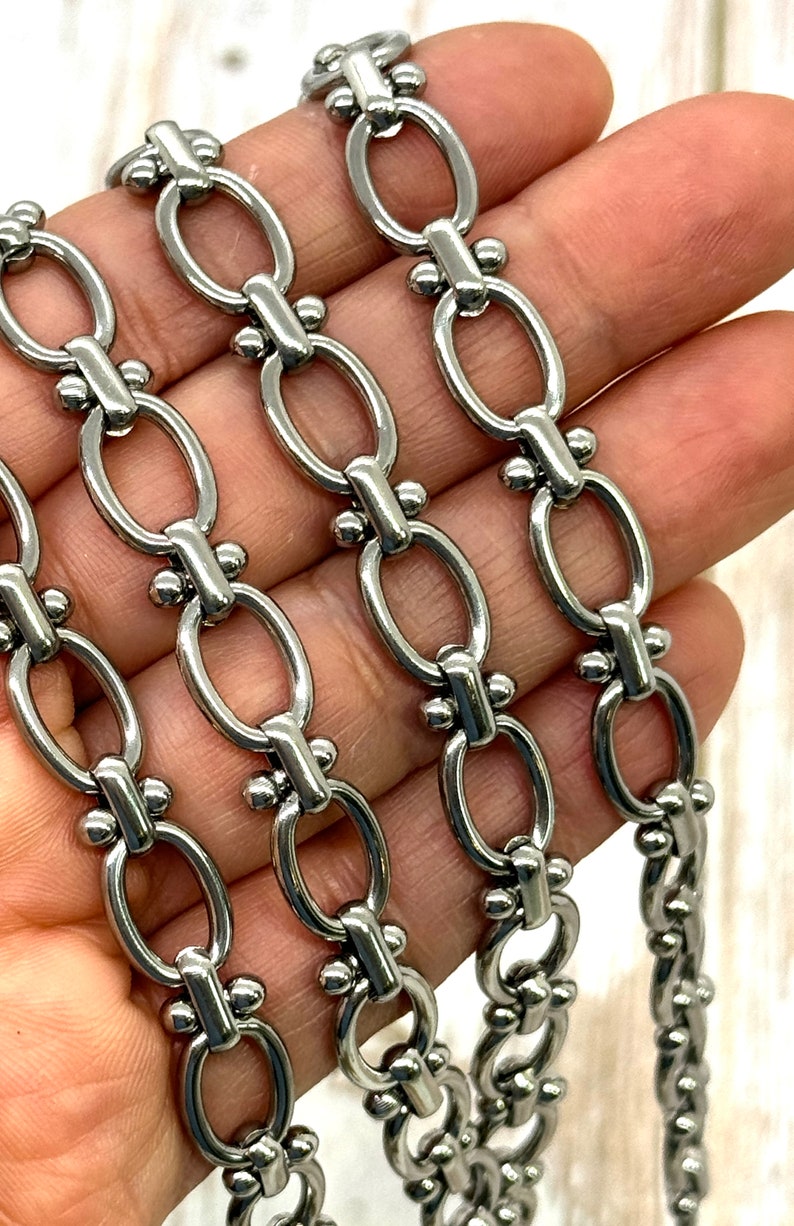 May include: Close-up of a silver-colored chain with oval links and decorative connectors. The chain is made of metal and has a polished finish. The links are connected by small, round accents.