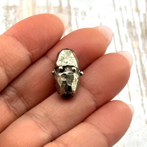 May include: A small, silver-toned metal bead with a faceted surface. The bead has a cylindrical shape with two small, round protrusions on each side. The bead is held in a hand.