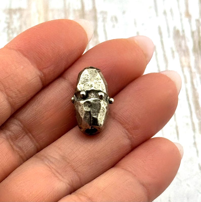 May include: A small, silver-toned metal bead with a faceted surface. The bead has a cylindrical shape with two small, round protrusions on each side. The bead is held in a hand.
