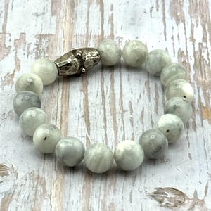 May include: A beaded bracelet featuring round, light gray beads with subtle variations in color. The bracelet is secured with a silver-toned clasp. The beads have a polished appearance, and the bracelet is displayed on a light-colored wooden surface.