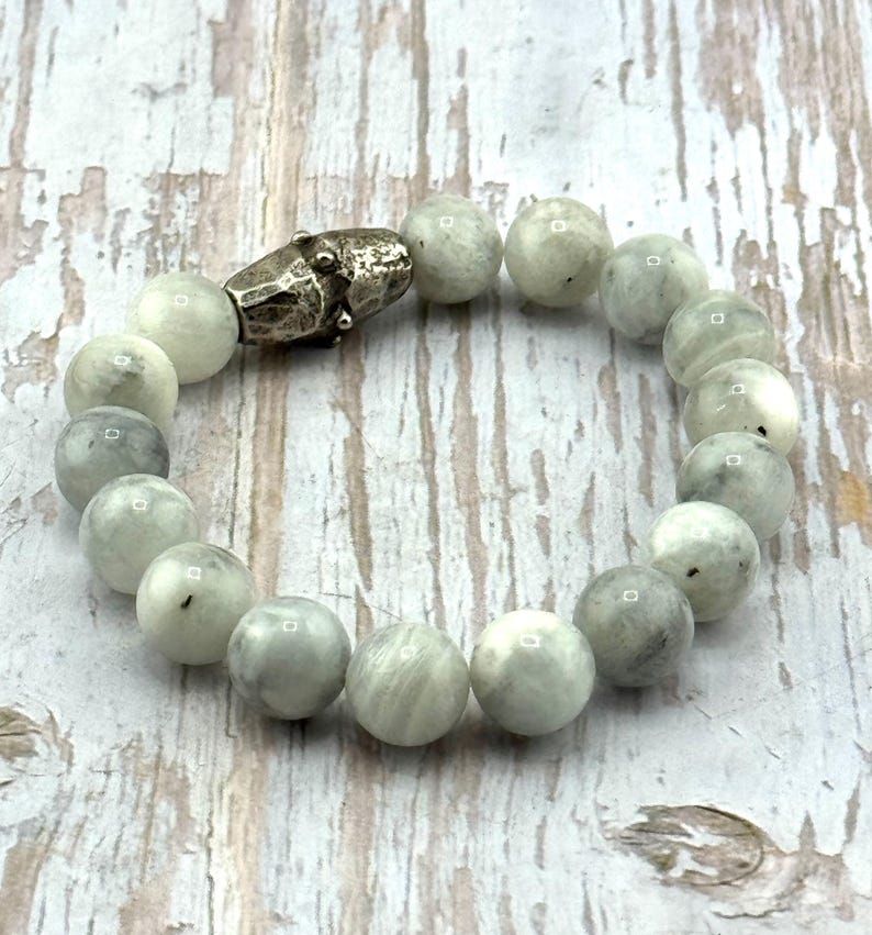May include: A beaded bracelet featuring round, light gray beads with subtle variations in color. The bracelet is secured with a silver-toned clasp. The beads have a polished appearance, and the bracelet is displayed on a light-colored wooden surface.
