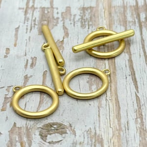 May include: Five gold-toned metal toggle clasps, including two round rings and three straight bars. The clasps are arranged on a light wood surface.