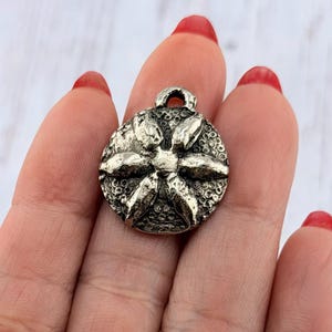 Op de afbeelding: Een zilverkleurige, ronde hanger met een bloemmotief. De hanger heeft een getextureerd oppervlak en een kleine lus aan de bovenkant om aan een ketting of koord te bevestigen. Het bloemmotief heeft zes bloemblaadjes en een gedetailleerd midden.