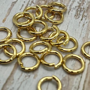May include: A pile of gold jump rings, a type of jewelry finding, are scattered on a white wooden surface. The rings are all the same size and are made of a shiny metal.