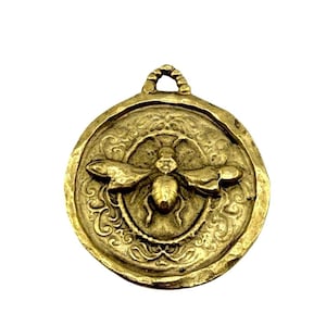 May include: A gold-colored pendant with a bee design. The round pendant features a detailed bee in the center, surrounded by an ornate border. The pendant has a loop at the top for hanging.