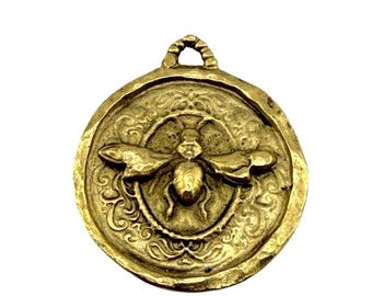 May include: A gold-coloured pendant with a bee design. The round pendant features a detailed bee in the centre, surrounded by an ornate border. The pendant has a loop at the top for hanging.