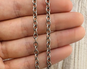 May include: A silver-toned metal chain necklace featuring a series of interconnected oval links. Each individual link displays a textured, ridged surface pattern that creates a subtle, repeating detail along the entire length of the chain.