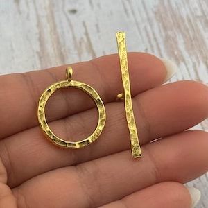 May include: A gold-colored toggle clasp set, featuring a textured, hammered finish. The set includes a circular ring and a straight bar with a small peg. Ideal for jewelry making and crafting.
