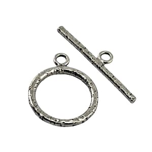 Peut inclure: Un fermoir toggle argenté composé d'un anneau circulaire et d'une barre droite. Les deux pièces présentent une finition de surface texturée et irrégulière. L'anneau possède une petite boucle sur le dessus et la barre une boucle centrée.