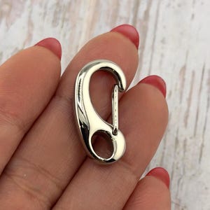 May include: A shiny, silver-colored carabiner clip. The clip has a curved design with a spring-loaded gate for secure closure. The metal has a polished, reflective surface.