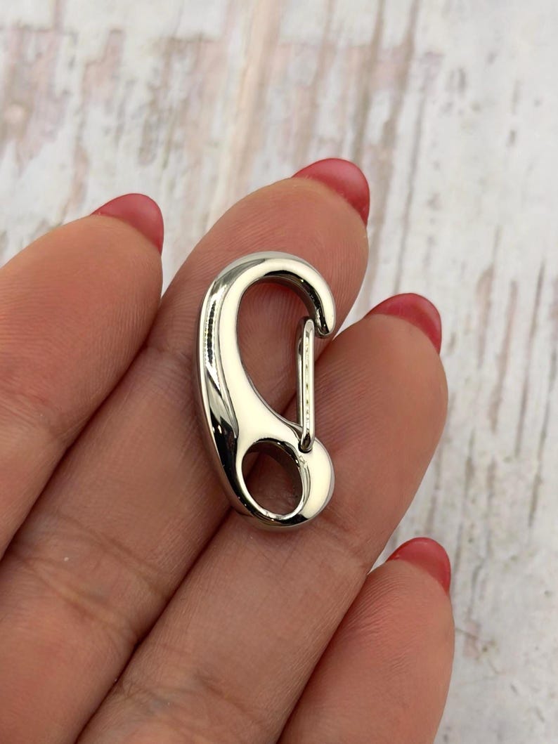 May include: A shiny, silver-colored carabiner clip. The clip has a curved design with a spring-loaded gate for secure closure. The metal has a polished, reflective surface.