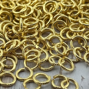 May include: Close-up of a pile of small, gold-colored jump rings. The rings have a textured, rope-like pattern around the outside. The rings are open and ready to be used for jewelry making or other crafts.