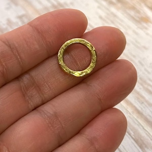 May include: A small, textured gold ring. The ring is circular with a slightly uneven surface, suggesting a handcrafted or vintage aesthetic. The ring is held in a person's hand, showcasing its size and detail.