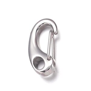 May include: A shiny, silver-colored carabiner clip. The clip has a rounded, oval shape with a spring-loaded gate. It features a small hole at the base and is designed for secure attachment. The carabiner is made of metal.