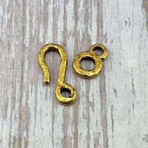 May include: Two small, gold-colored metal clasps. One is a hook shape, and the other is a circular shape with a hole in the center. The clasps have a textured surface and are likely for jewelry or crafts.